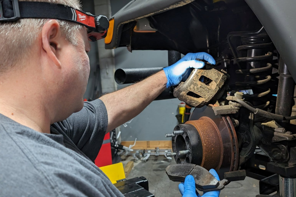 brake repair, auto repair shop in summerville, SC. SBS Automotive. Technician at S.B.S Automotive performing a brake service, holding worn brake pads and a caliper while inspecting a rusted rotor on a vehicle’s suspension system.