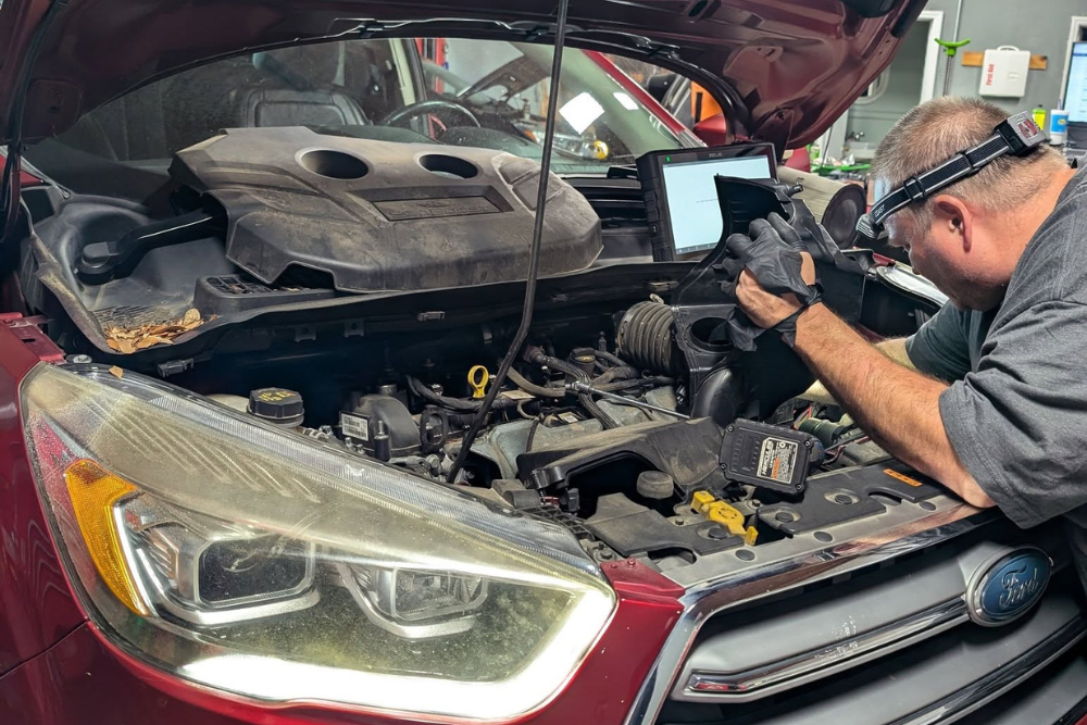 Car maintenance in Summerville, SC. SBS Automotive Maintenance and Repair. A mechanic wearing gloves and a headlamp inspects the engine of a red Ford SUV with the hood open, using a diagnostic tablet in an auto repair shop.