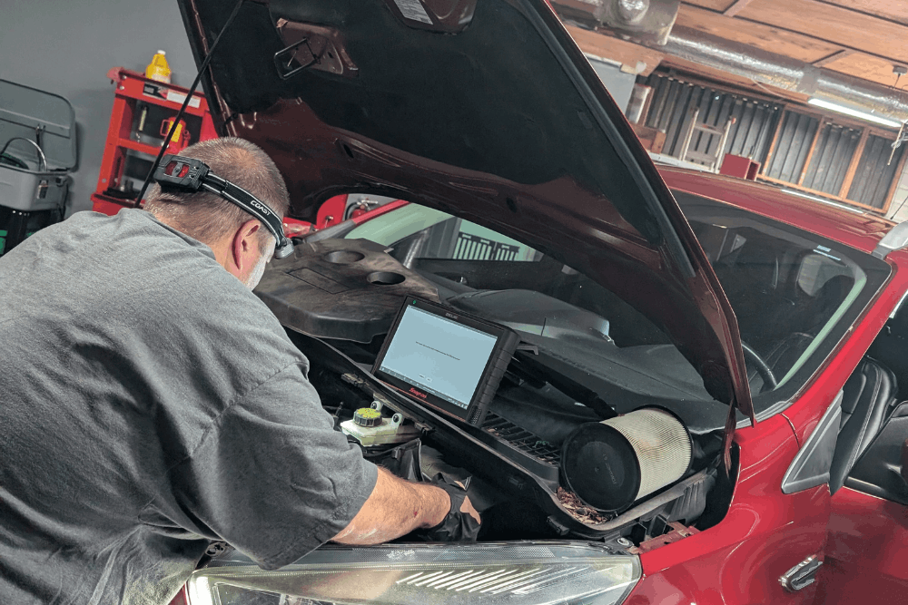 Car inspection, auto repair in Summerville, SC by SBS Automotive Maintenance and Repair. Image of a technician scanning a red vehicle under the hood using a diagnostic tablet to identify issues and restore performance.