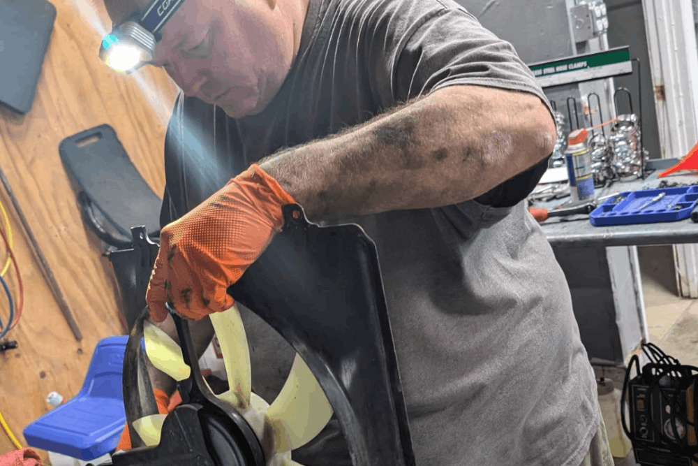 Stop Leak Product Dangers for Car A/C Systems - SBS Automotive -Man wearing a headlamp and orange gloves working on a car fan in a garage, surrounded by tools, with a focused and dedicated expression.